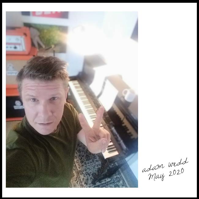 Adam Wedd gives the V peace sign to the camera, he's standing in front of a piano