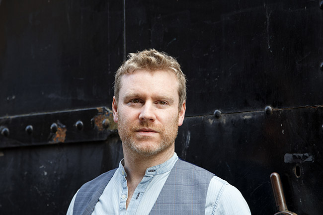 A promotional shot of Fiach Moriarty, looking pensive and into the distance, he wears a blue shirt and waistcoat